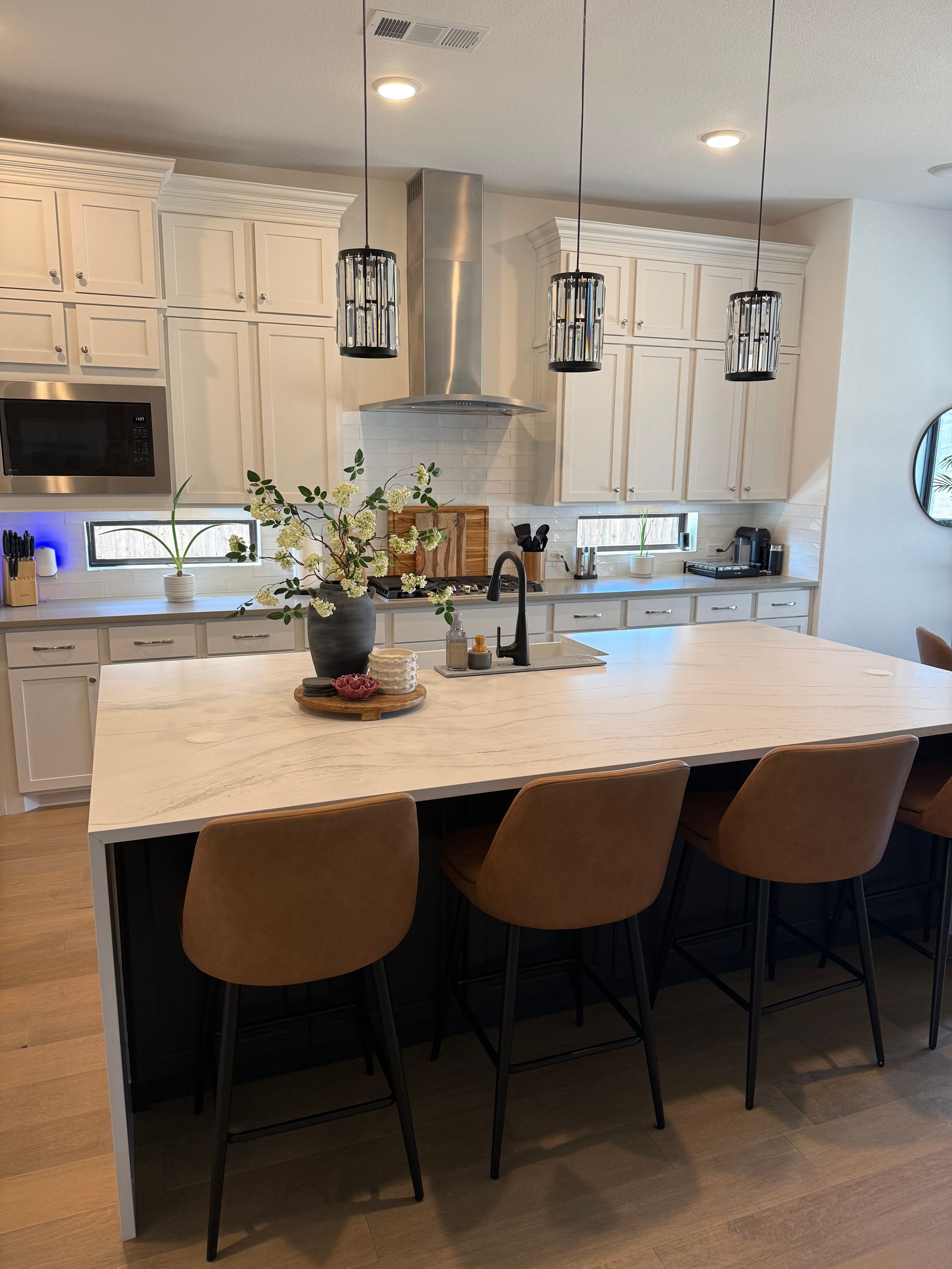 A modern kitchen featuring white cabinets, a large marble-topped island with three tan stools, and hanging pendant lights.