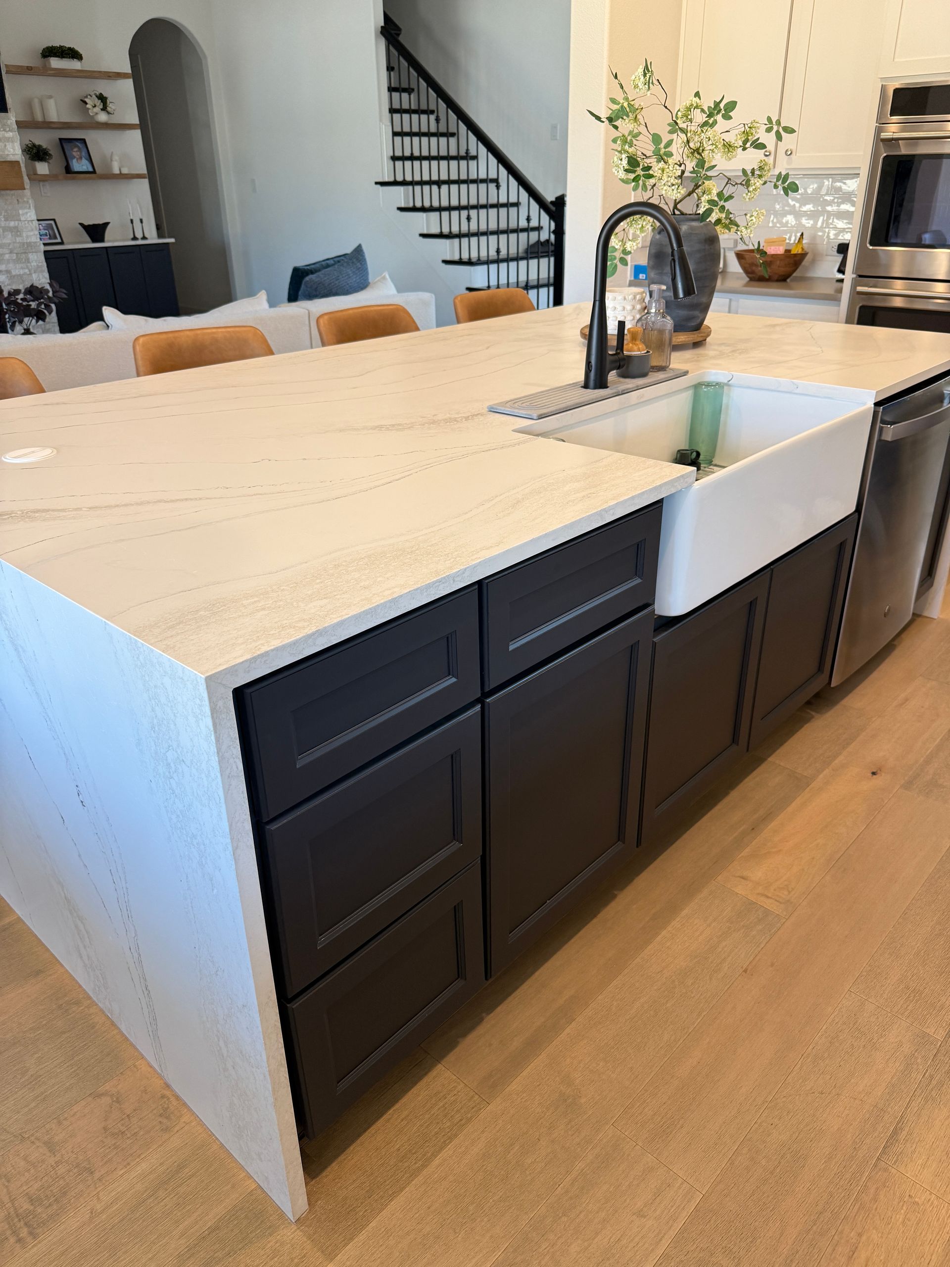A kitchen island with dark cabinetry, a white farmhouse sink, and a light-colored stone countertop on wood flooring.