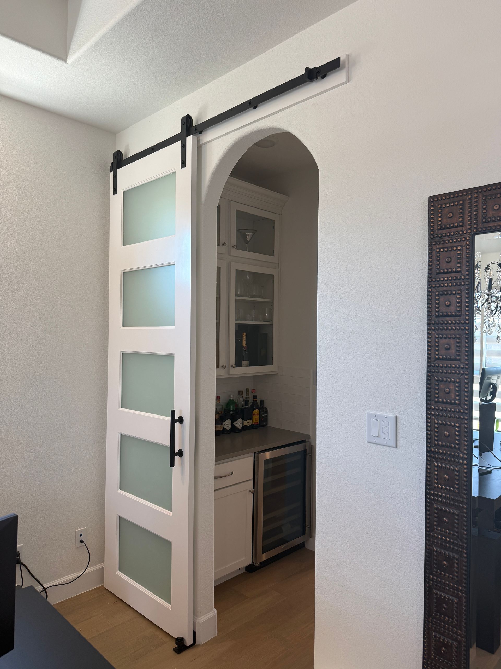 A white barn door with five frosted glass panels slides on a black track to close off an arched doorway to a home bar.