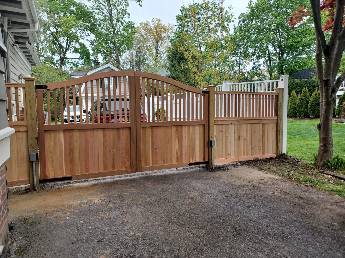 A black metal gate framed by brick pillars and stone walls in front of a house on a tree-lined, sunny street.