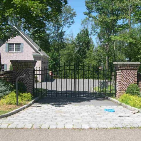 Black metal gate framed by decorative brick pillars at the entrance of a paved driveway with a house in the background.