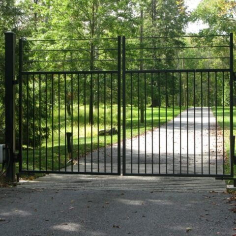 A black metal double-swing gate stands closed across a gravel driveway, flanked by green trees and foliage.