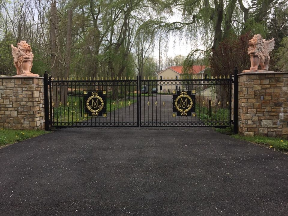 A black iron driveway gate with gold emblems, flanked by stone pillars topped with winged lion statues on a tree-lined lot.