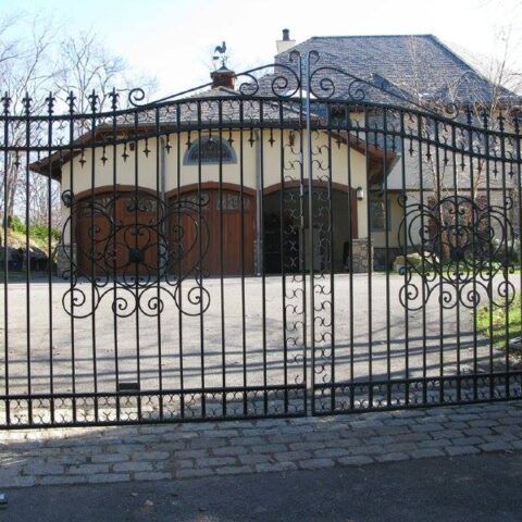A tall, arched black wrought iron gate stands in front of a house with a three-car garage and a stone driveway.