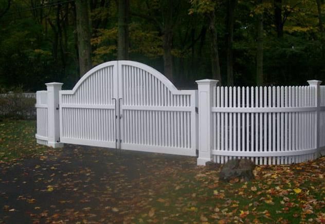 A white, arched double gate with vertical pickets, attached to a matching picket fence in a wooded area with fallen leaves.