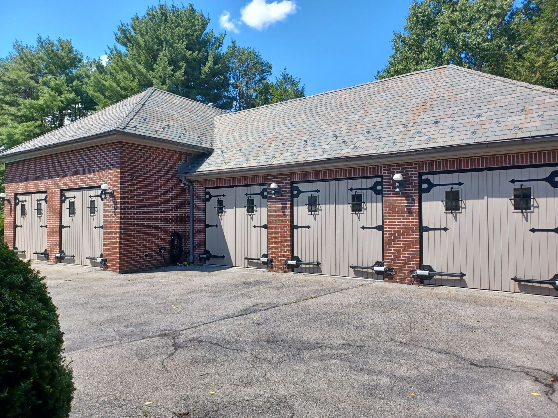 A red brick building with four light-colored carriage-style garage doors, a shingled roof, and a gravel driveway.