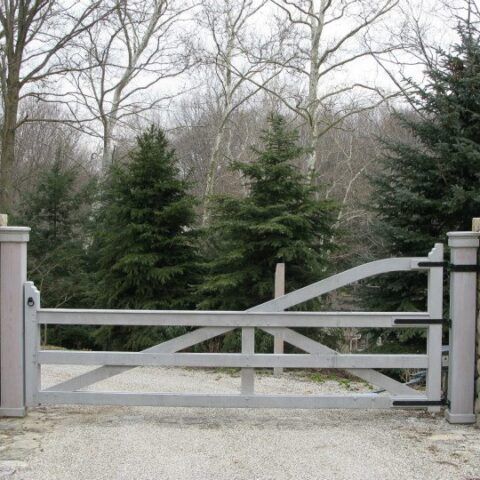 A light-colored wooden gate with a diagonal brace stands on a gravel driveway between stone pillars, set against trees.