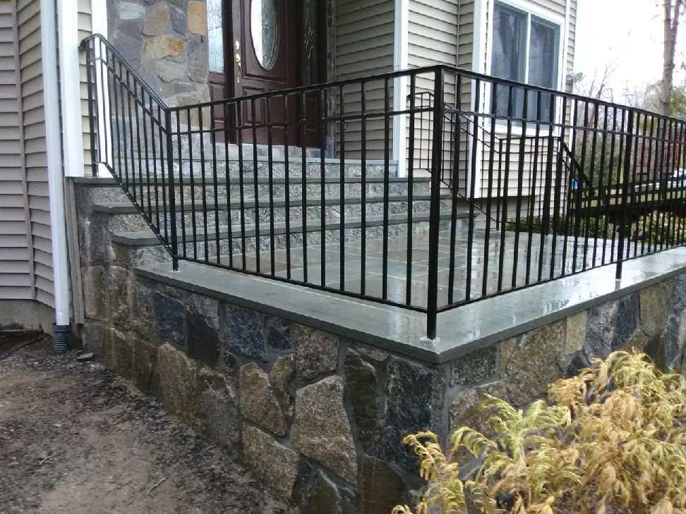 Stone porch steps with a black metal railing leading to a home entrance with brown siding and a wooden front door.