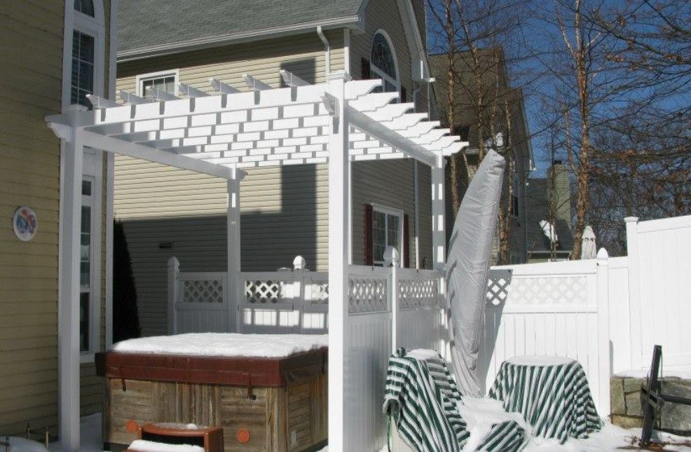 A snowy backyard scene featuring a white pergola over a hot tub, next to a white privacy fence and covered outdoor chairs.