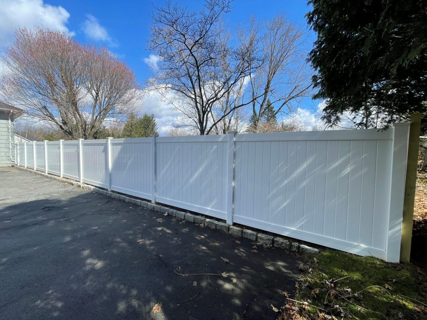 A white vinyl privacy fence lines a paved driveway, with bare trees and a blue sky in the background.