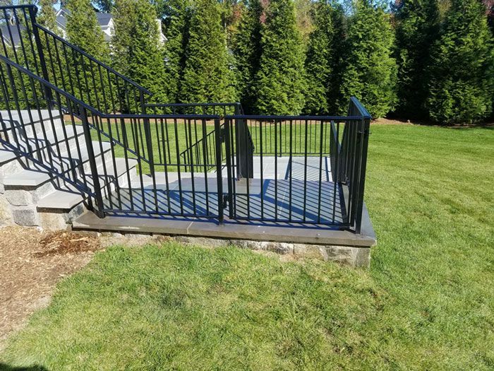 A black horizontal-slat gate stands across a gravel driveway in front of a white house on a cloudy day.