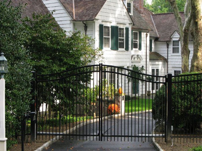 Black metal driveway gate in front of a white house with green shutters surrounded by trees and shrubs.