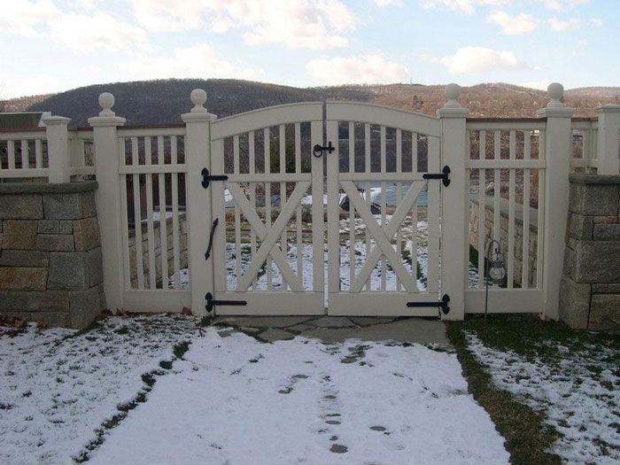 A white vinyl privacy fence installed atop a tiered stone retaining wall against a blue sky and leafless trees.