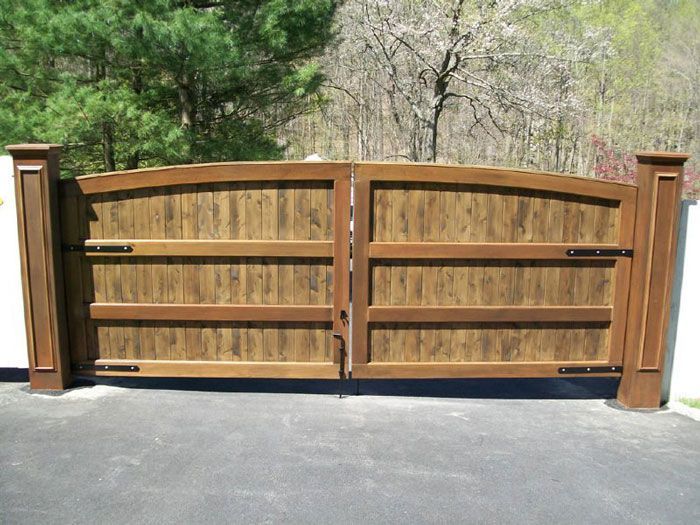 A double wooden gate with an arched top, mounted between two sturdy pillars against a backdrop of trees.