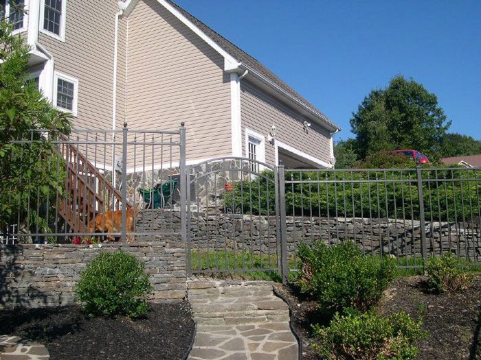 A black metal fence sits atop a tan stone wall in front of a Tudor-style building on a paved patio.