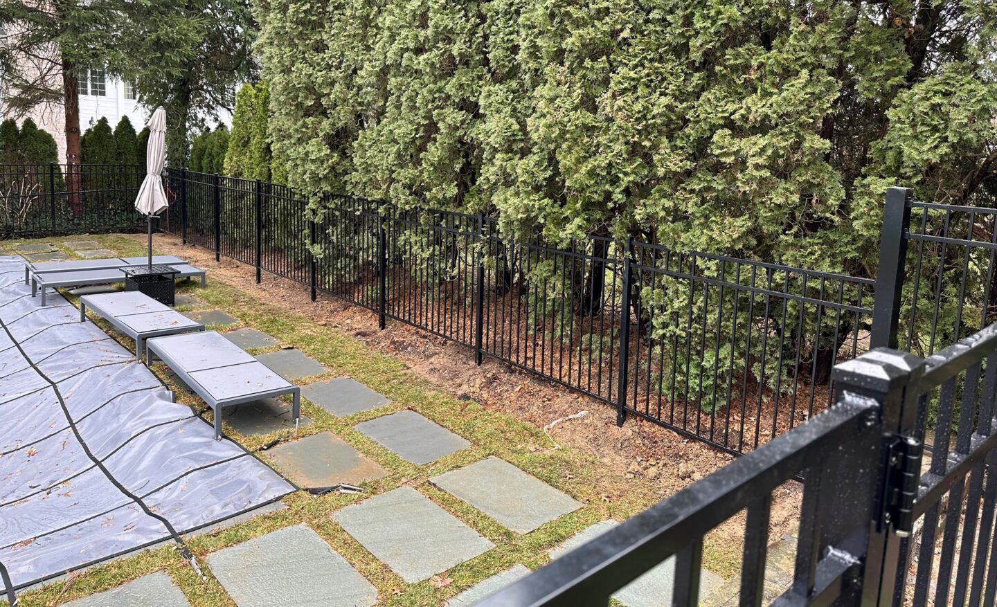A brown privacy fence sits atop a stone retaining wall next to a small creek, with a building and car in the background.