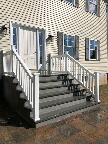 A covered swimming pool with a stone patio, enclosed by a black metal fence, behind a house with a wooden deck.