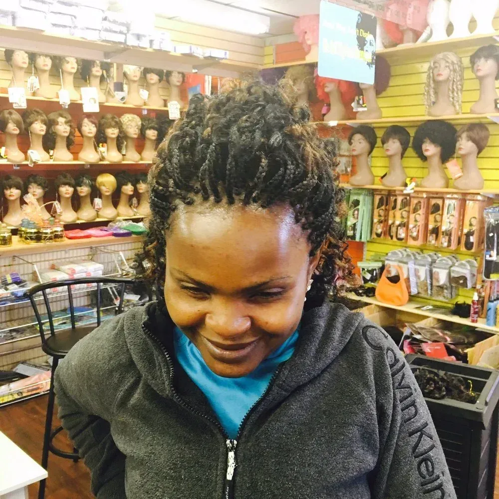 Woman in a wig store with braided hair, smiling.  Wigs on display in the background.