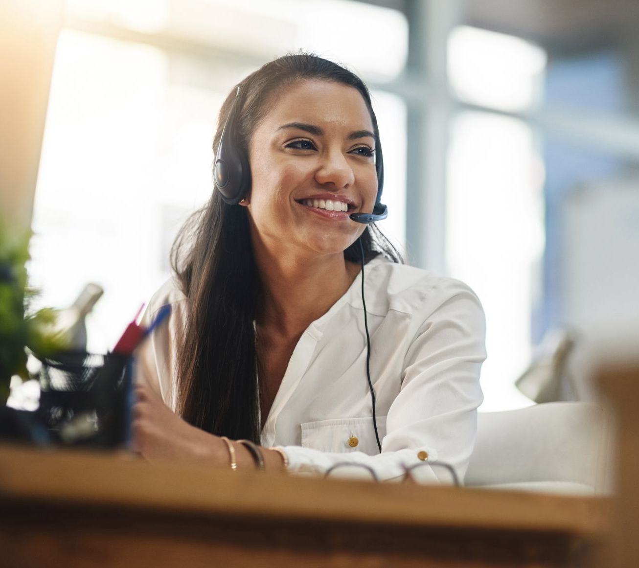 Woman wearing headset, smiling while working at a desk in a bright office environment.