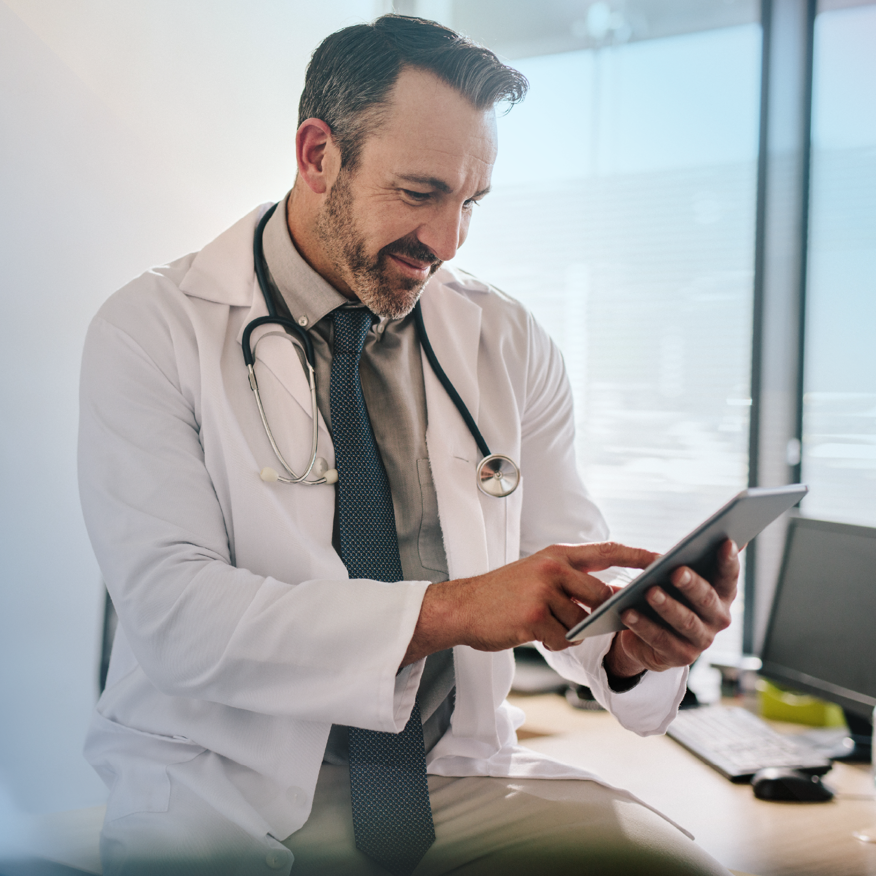 Doctor in white coat using a tablet, smiling, stethoscope around neck, office setting.