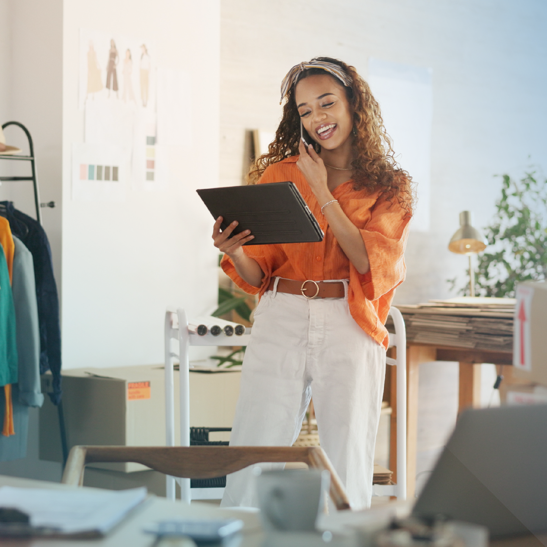 Woman talking on phone, holding tablet, wearing orange shirt and white pants in a brightly lit room with office supplies.