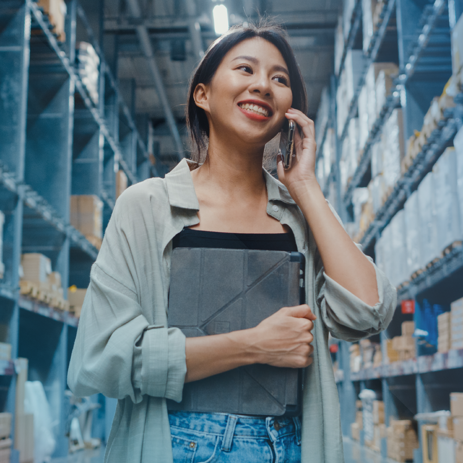 Woman in warehouse holding tablet, smiling while on the phone, shelves of packages in background.