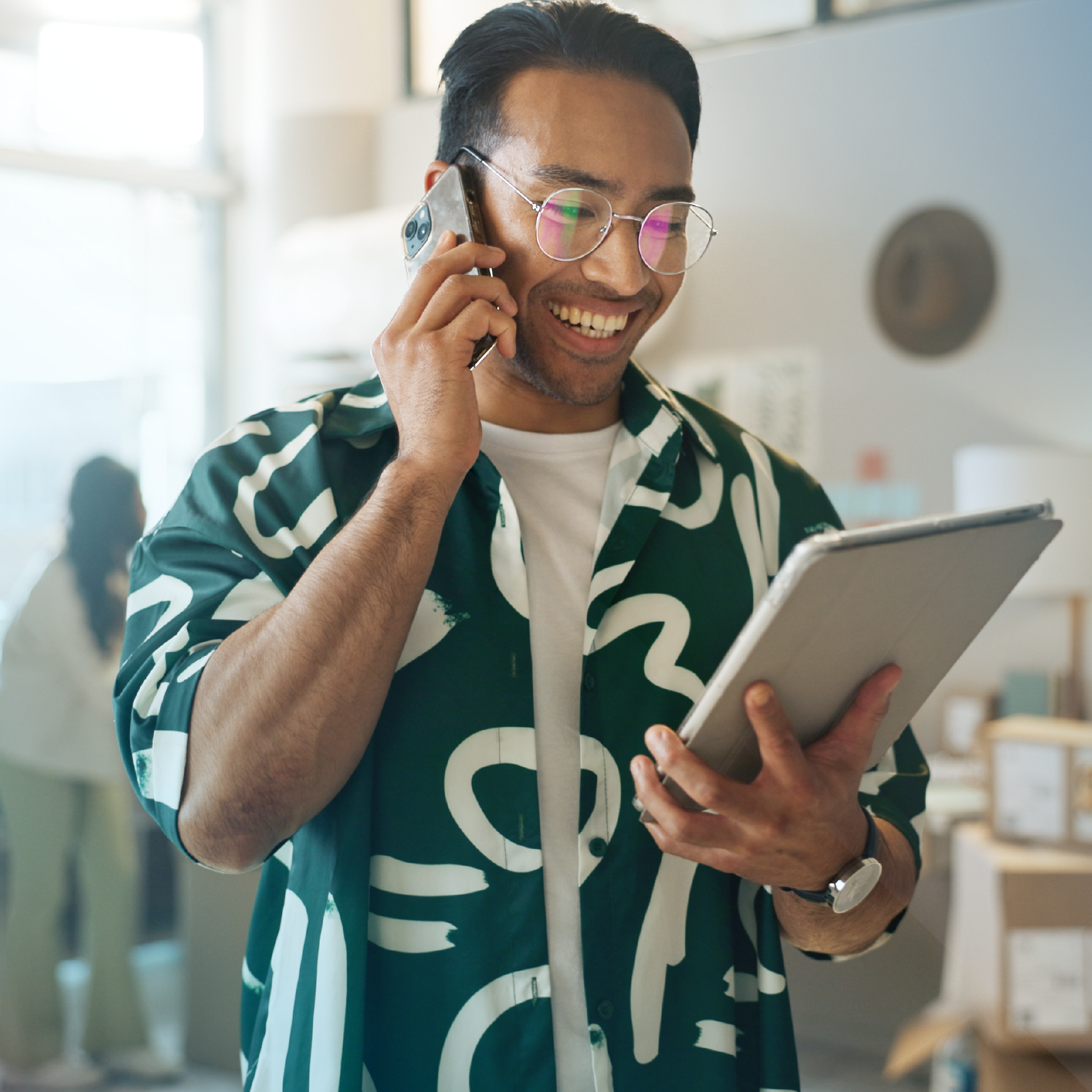 Man on phone, smiling, looking at tablet in a brightly lit office. Wearing glasses, green patterned shirt.
