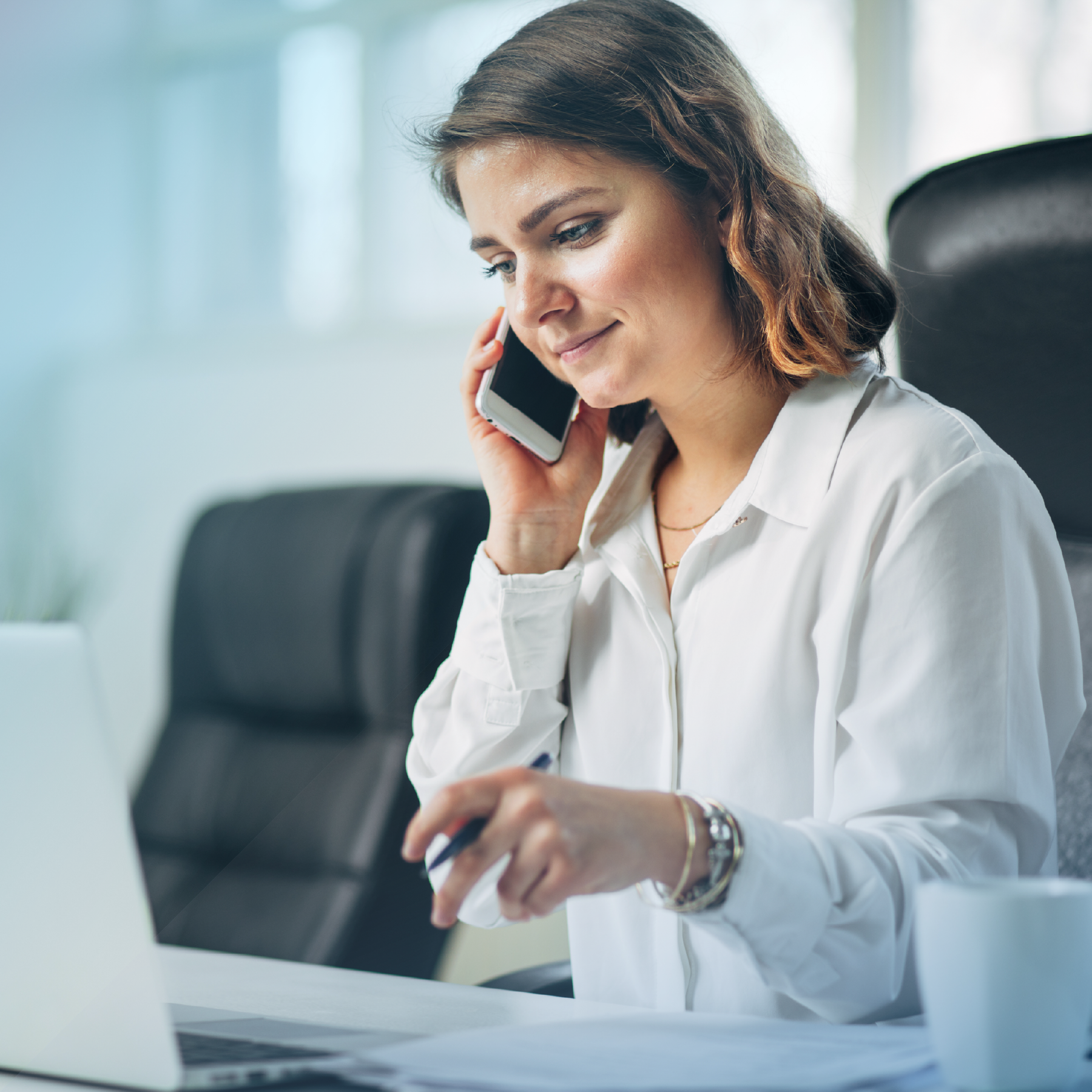 Woman in white shirt on phone, working at desk with laptop and coffee cup.
