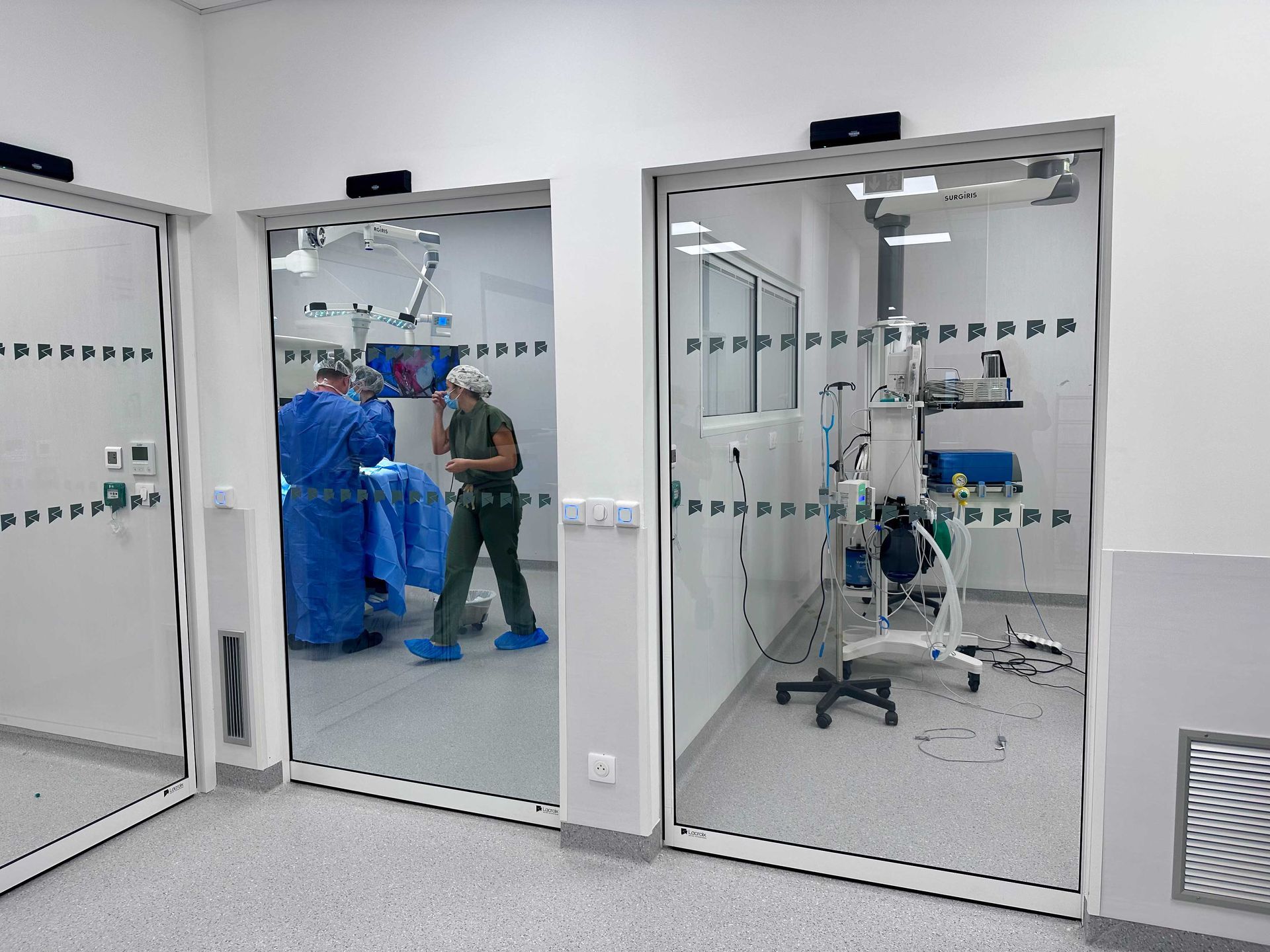 Trois portes vitrées automatiques donnent accès aux salles d'opération ; l'une montre le personnel médical au travail, l'autre présente du matériel.