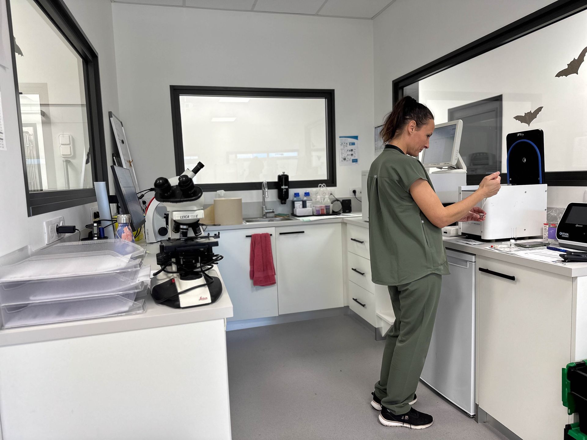 Une femme en blouse médicale travaille dans un laboratoire. Microscope et matériel de laboratoire sont posés sur les paillasses. Pièce bien éclairée.