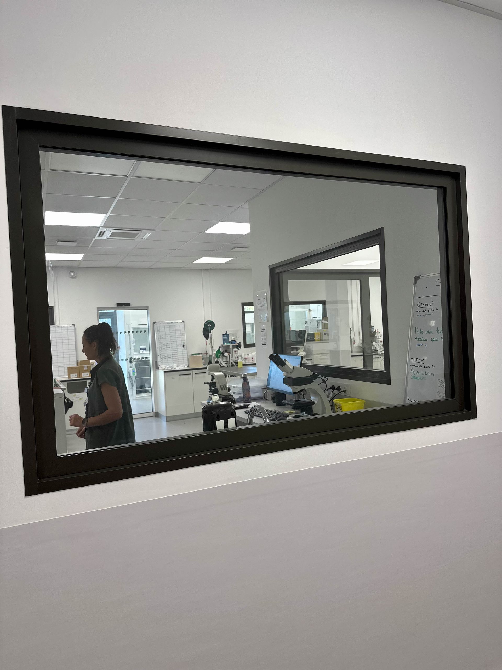 Une fenêtre rectangulaire donne sur l'intérieur d'un laboratoire. Une personne se tient près d'un appareil. Cadre de fenêtre noir sur un mur blanc.