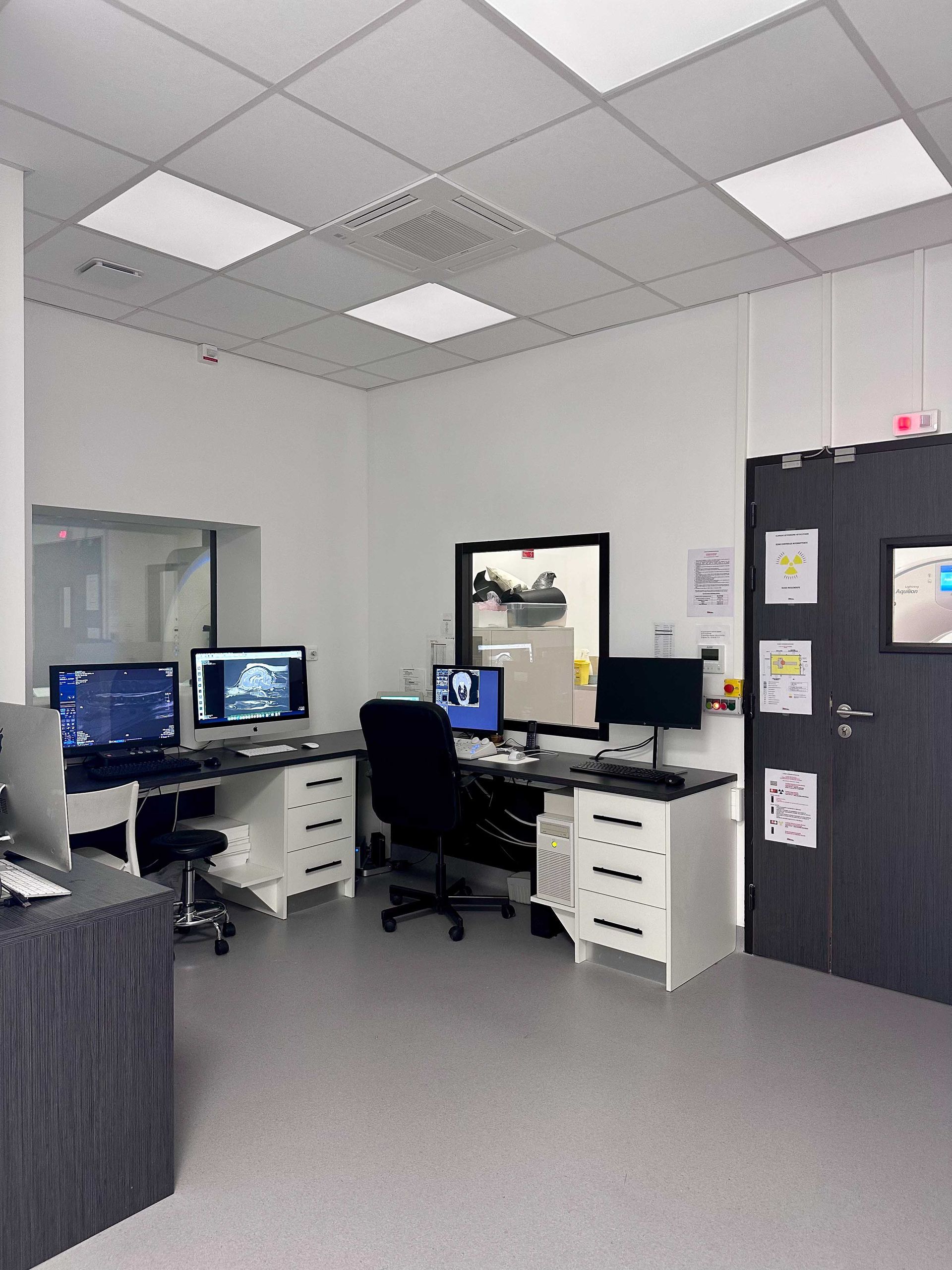 Bureau avec ordinateurs, bureaux et porte noire. Murs blancs, sol gris et éclairage zénithal.
