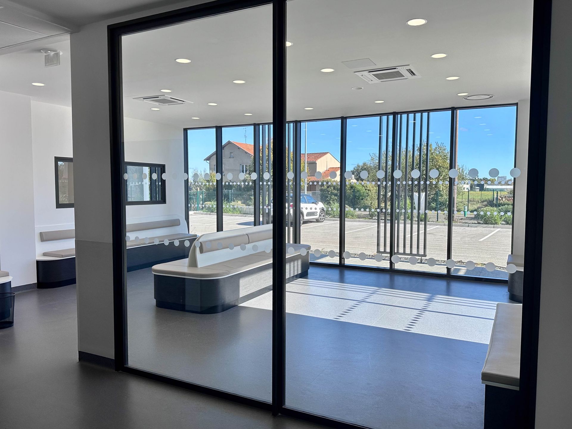 Salle d'attente moderne avec parois vitrées, bancs et vue sur l'extérieur.