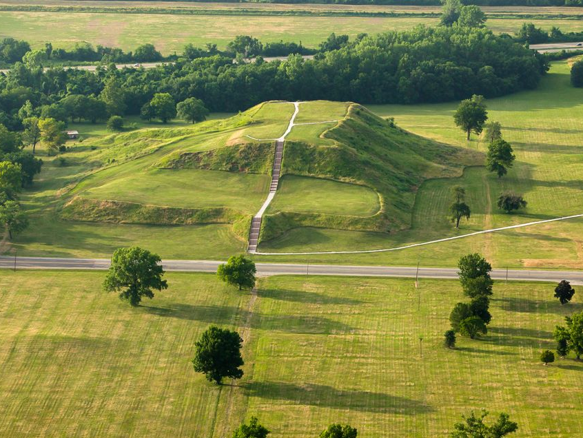 Aerial view of a large, grassy Native American mound in the landscape, with a road and trees nearby.
