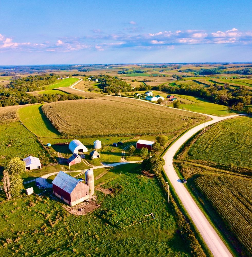 Aerial view of a rural farm with fields, barns, a silo, and a road winding through the landscape under a blue sky.