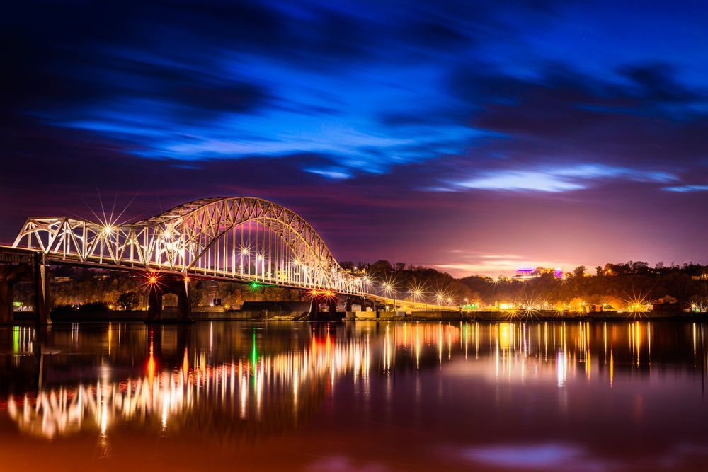 Night view of illuminated arched bridge over a still body of water, with vibrant reflections in the water.