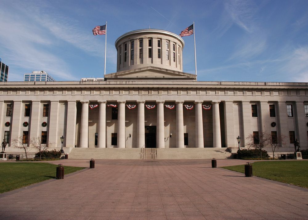 Ohio Statehouse, Columbus, white neoclassical building with columns, flags, and a domed roof under a blue sky.