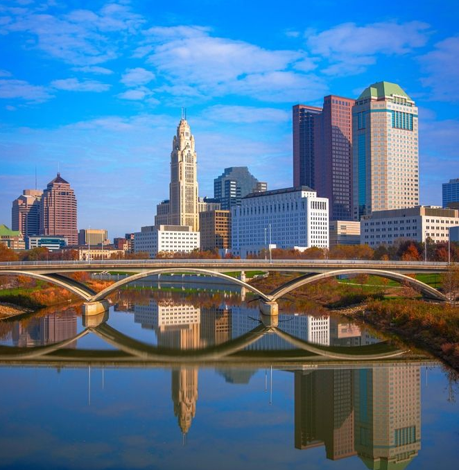 Columbus, Ohio skyline reflected in the water under a blue sky, with a bridge in the foreground.