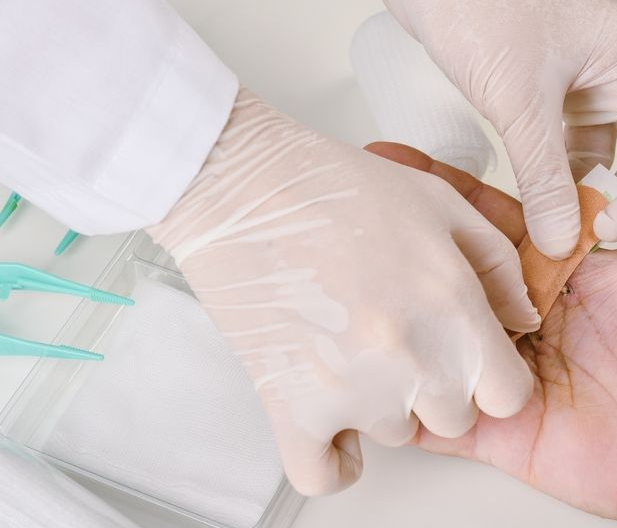 Gloved medical hands checking a patient’s hand pulse on a white clinic bed