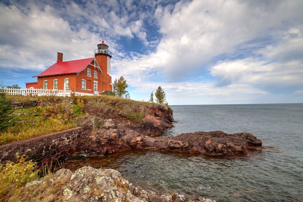 Lighthouse with red roof on rocky shore under blue sky and clouds.
