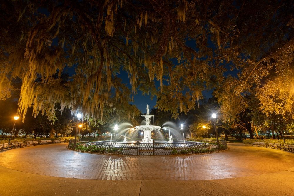 Fountain in a park at night, lit by streetlights and surrounded by trees with Spanish moss.