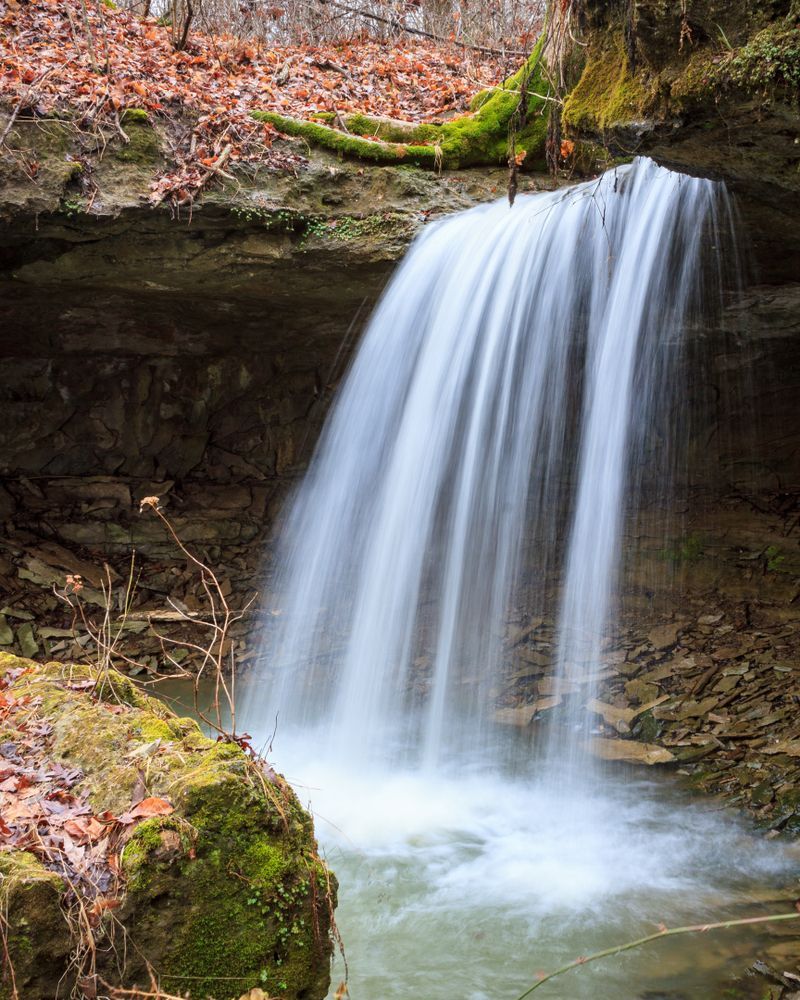 Waterfall cascading from a rock overhang into a pool below, surrounded by moss and autumn leaves.