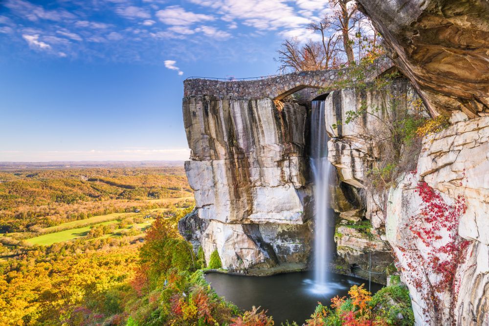 Waterfall cascading over a cliff edge with a stone bridge above. Lush autumn foliage surrounds.
