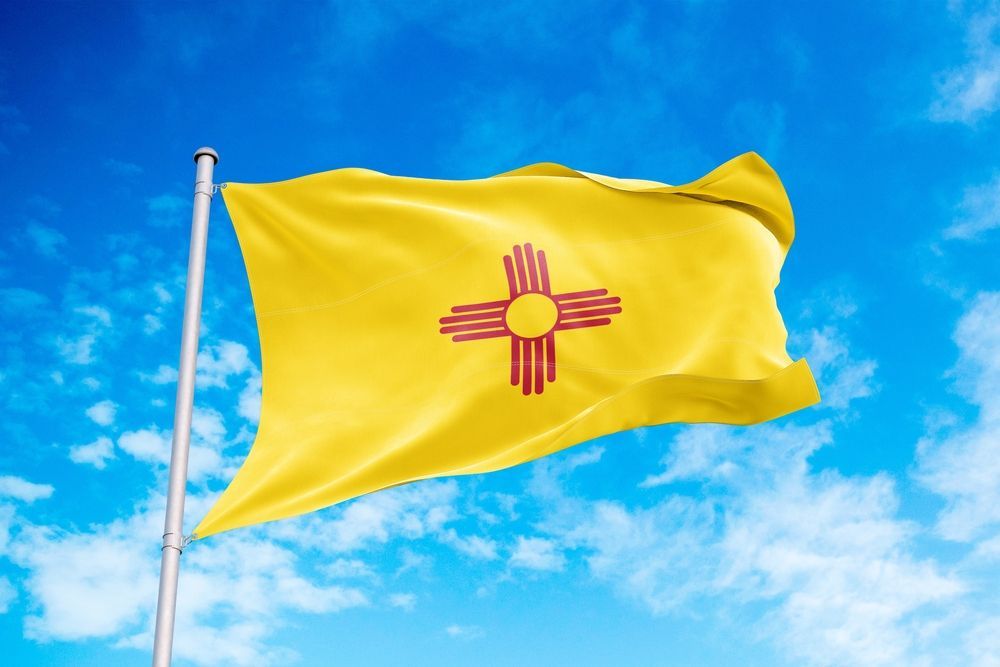 New Mexico state flag waving in the wind: a yellow field with a red Zia symbol against a blue sky.
