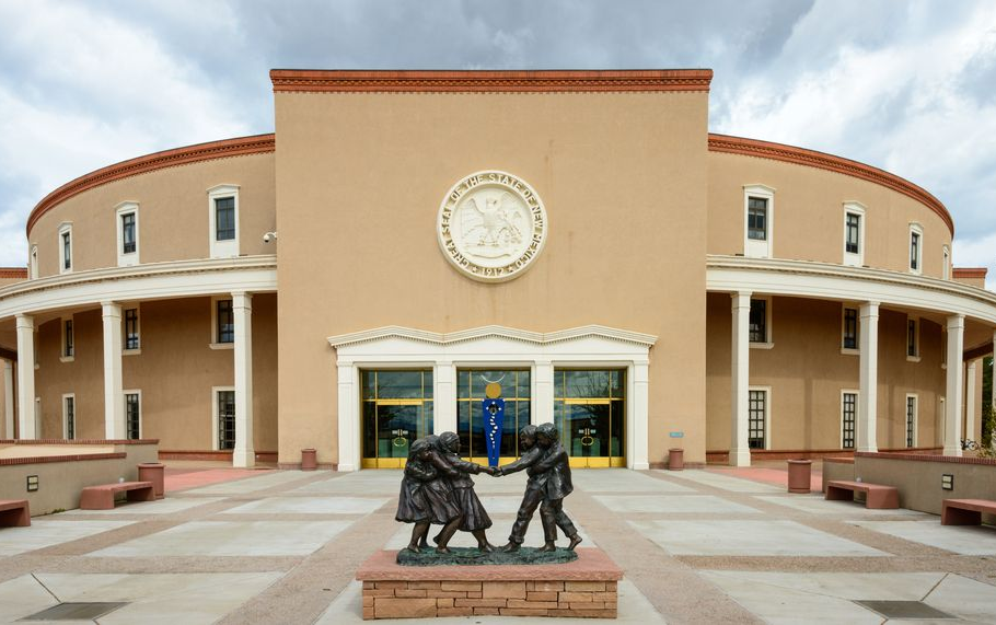 New Mexico State Capitol building, tan adobe with columns and central circular design. Bronze statue in front.