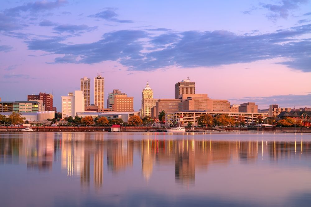 City skyline reflecting in calm water at sunset; buildings silhouetted against pink and purple sky.