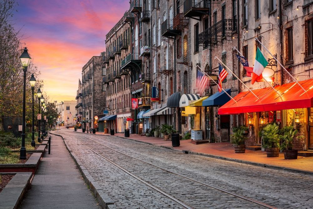 Cobblestone street in Savannah, Georgia, lined with historic buildings, colorful awnings, and flags, at sunset.