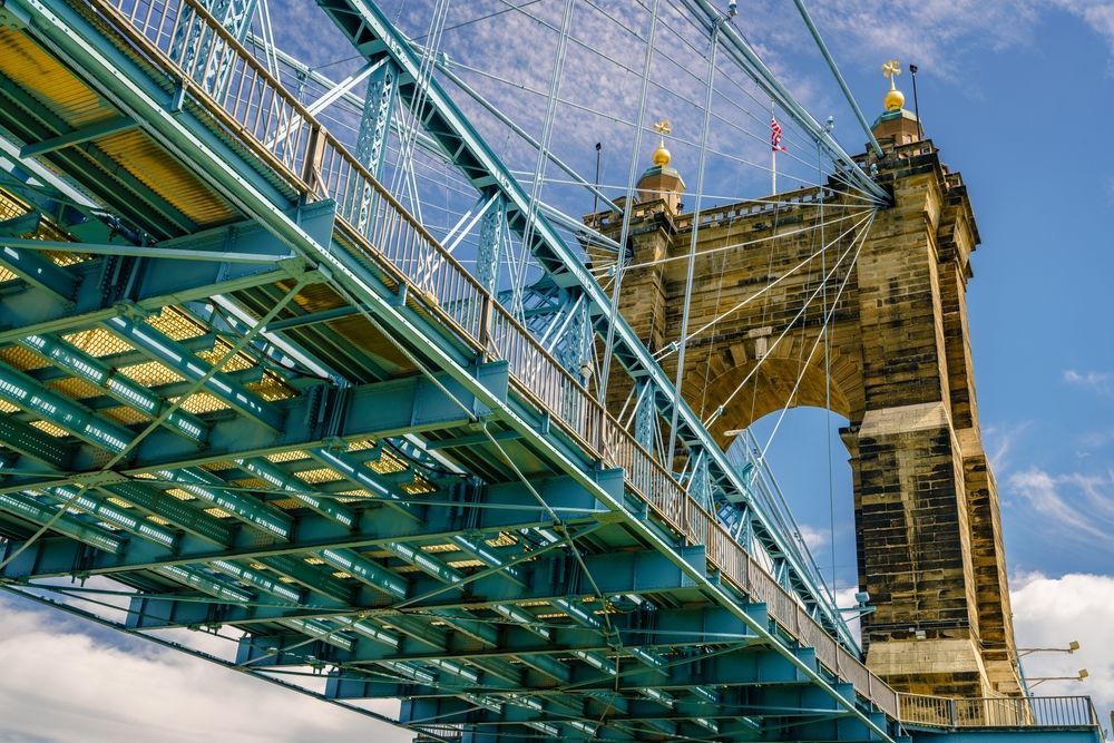 Blue and turquoise suspension bridge towers against a blue sky; stone tower with arches.