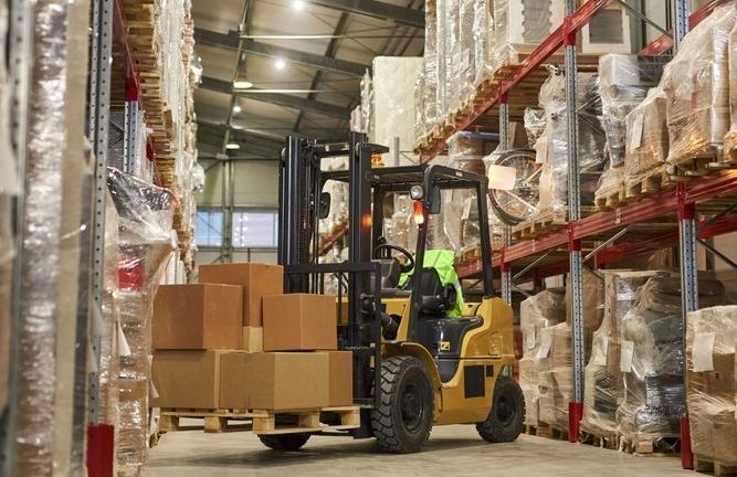 A man is driving a forklift in a warehouse filled with boxes.