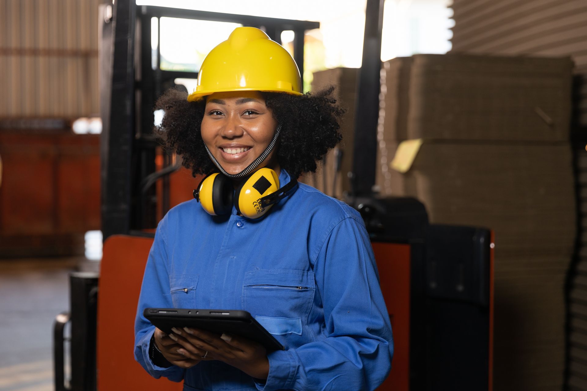 A woman wearing a hard hat and earmuffs is holding a tablet in a warehouse. she is reading up on the Sales Tax Exemption for Manufacturers